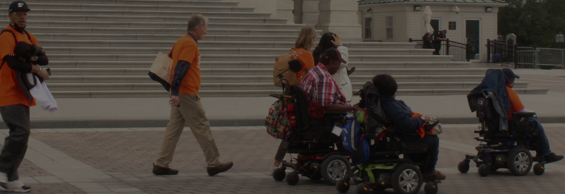 A group of disability rights advocates, some using wheelchairs and others walking, move together outside a government building with large stone steps in the background. They are wearing orange shirts and carrying bags, showing solidarity and engagement in advocacy efforts.